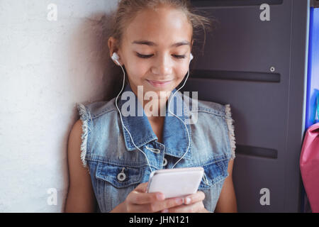 Elementary schoolgirl listening music through headphones while using mobile phone by wall at school Stock Photo