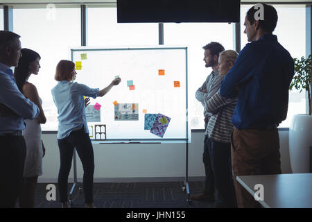 Businesswoman sticking adhesive notes on whiteboard while colleagues standing with arms crossed at office Stock Photo