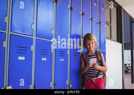 Schoolboy using mobile phone while standing by lockers in corridor at school Stock Photo