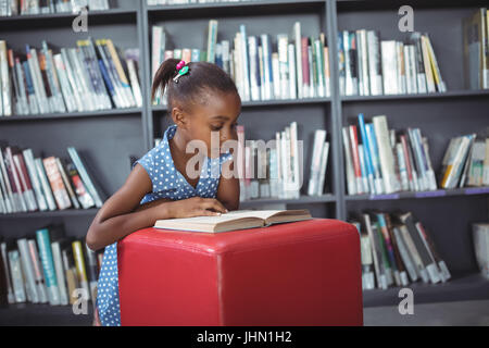 Girl reading book on ottoman against bookshelf in library Stock Photo
