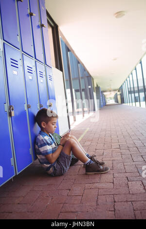 Side view of boy using mobile phone while sitting by lockers in corridor at school Stock Photo