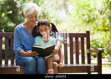 Smiling grandmother reading novel to granddaughter sitting on wooden bench at garden Stock Photo