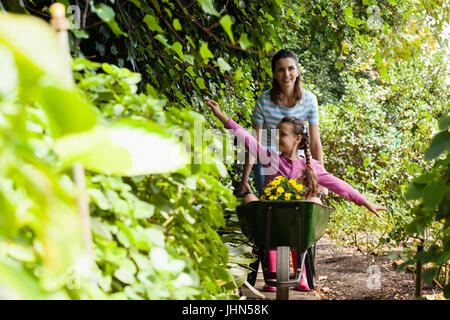 Smiling woman pushing girl sitting with outstretched in wheelbarrow on footpath amidst plants at garden Stock Photo