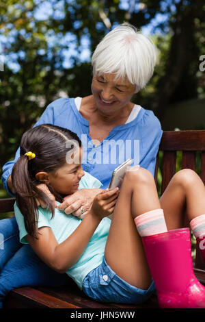 Cheerful senior woman enjoying with girl holding mobile phone on bench at backyard Stock Photo