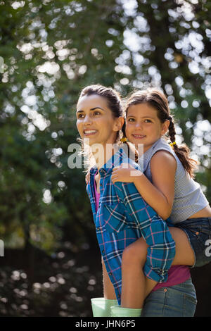 Thoughtful smiling mother piggybacking daughter against trees in backyard Stock Photo