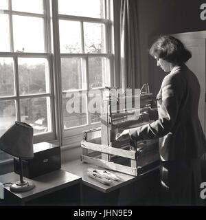 1950s, historical, picture shows a young lady standing by a sash window using a traditional small wooden table-top hand-weaving machine or loom tp produce a piece of cloth or fabric, England, UK. Stock Photo