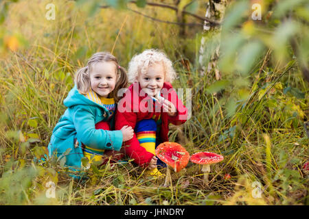 Happy laughing kids play in beautiful sunny autumn park. Little boy and girl watching toadstool mushroom in fall forest. Children hiking and playing o Stock Photo