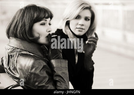 Two happy young fashion women smoking on the bench Stock Photo