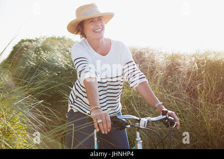 Smiling mature woman riding bicycle on sunny beach grass path Stock Photo