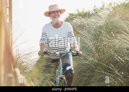 Smiling mature woman riding bicycle along beach grass Stock Photo