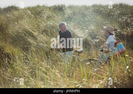 Mature couple walking bicycles in sunny beach grass Stock Photo