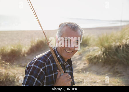 Portrait smiling senior man with fishing rod walking on sunny beach Stock Photo