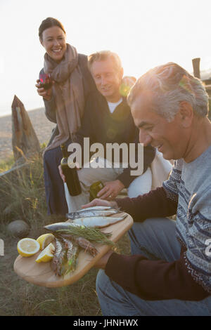 Mature friends drinking wine and barbecuing fish on sunset beach Stock Photo