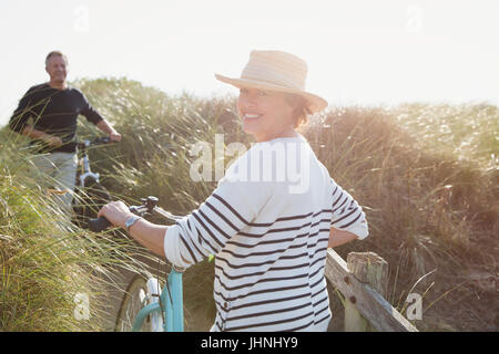 Portrait smiling mature woman walking bicycle on sunny beach grass path Stock Photo