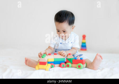 Adorable Asian baby boy 9 months sitting on bed and playing with color wooden train toys at home. Stock Photo