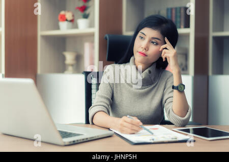 Portrait of a beautiful Asian young businesswoman sitting in the office, holding document and thinking. Stock Photo