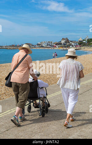 Couple pushing elderly woman in wheelchair along promenade seafront at Lyme Regis, Dorset in July Stock Photo