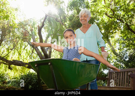 Smiling senior woman pushing granddaughter sitting in wheelbarrow at backyard Stock Photo