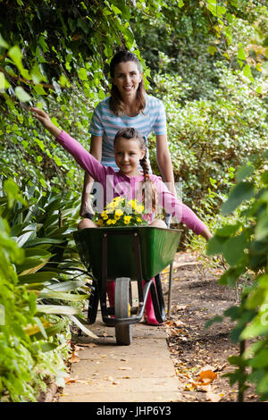 Woman pushing girl sitting with outstretched in wheelbarrow on footpath amidst plants at garden Stock Photo