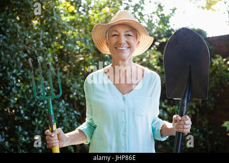 Portrait of smiling senior woman holding garden fork and shovel at backyard Stock Photo