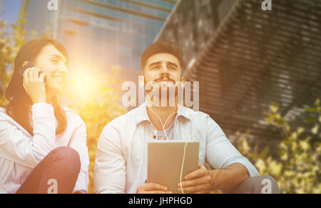 Man listening music with headphone while woman is using her mobile phone  against low angle view of modern buildings Stock Photo