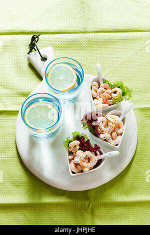 Prawn cocktail  in serving bowls in the form of hearts on a gray background. On a marble cutting board. Two glasses of water with ice Stock Photo