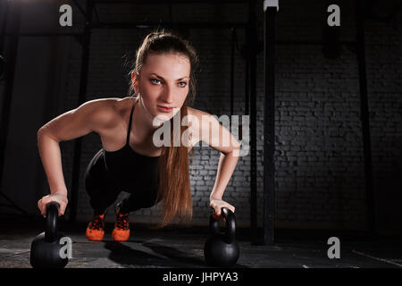 Young athlete woman in the black sportwear doing push ups on two kettlebells against brick wall in the gym. Stock Photo