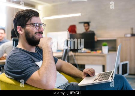 Photo bearded banker working in a modern office overtime. Man sitting in yellow chair at night. Using contemporary laptop, blurred background. Soft li Stock Photo