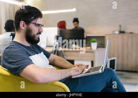 Photo bearded banker working in a modern office overtime. Man sitting in yellow chair at night. Using contemporary laptop, blurred background. Soft li Stock Photo