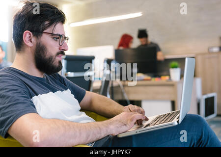 Photo bearded banker working in a modern office overtime. Man sitting in yellow chair at night. Using contemporary laptop, blurred background. Soft li Stock Photo