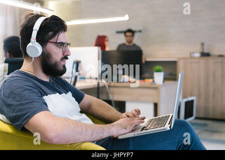 Photo bearded banker working in a modern office overtime. Man sitting in yellow chair at night. Using contemporary laptop, blurred background. Soft li Stock Photo