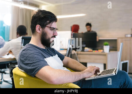 Photo bearded banker working in a modern office overtime. Man sitting in yellow chair at night. Using contemporary laptop, blurred background. Soft li Stock Photo