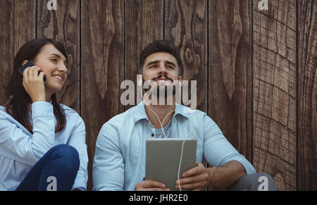 Man listening music with headphone while woman is using her mobile phone  against wood panelling Stock Photo