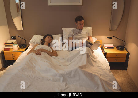 Man reading book while woman resting on bed at home Stock Photo