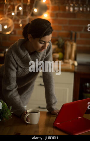 Young woman using laptop at table while standing in kitchen Stock Photo