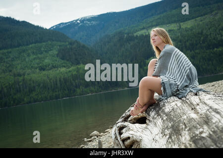 Side view of young woman sitting on log at lakeshore Stock Photo