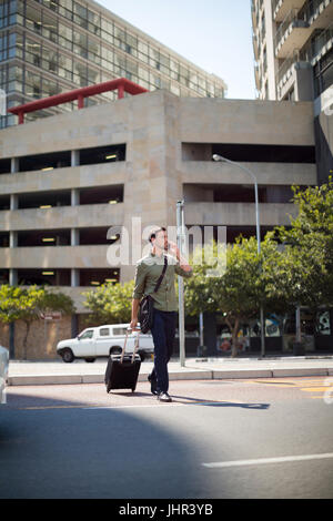 Man walking with suitcase while talking on mobile phone against buildings Stock Photo