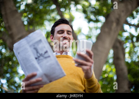 Smiling man with newspaper using mobile phone in park Stock Photo
