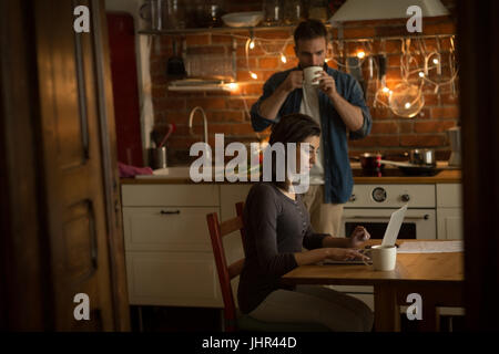 Man having drink while woman using laptop at table in kitchen Stock Photo