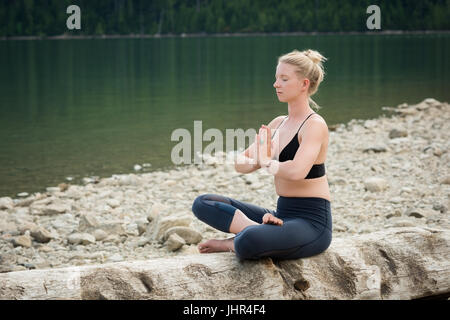 Young woman meditating while practicing yoga on log at lakeshore Stock Photo