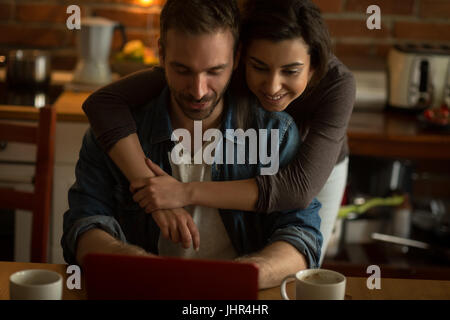 Woman embracing man using laptop at table in kitchen Stock Photo
