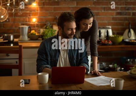Couple reading document while using laptop at table in kitchen Stock Photo