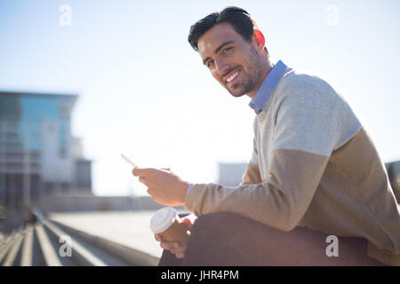 Portrait of smiling businessman sitting on staircase with mobile phone and coffee Stock Photo