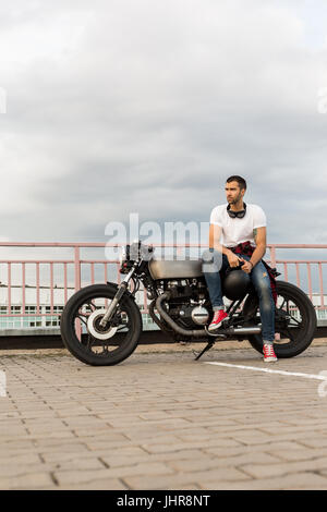 Rider guy with beard and mustache in red sneakers and white t-shirt sit on classic style biker cafe racer motorcycle. Bike custom made in vintage gara Stock Photo