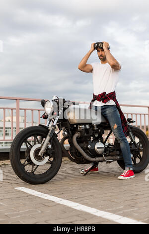 Handsome rider guy with beard and mustache posing while put on protection glasses near classic style biker cafe racer motorcycle. Bike custom made in  Stock Photo