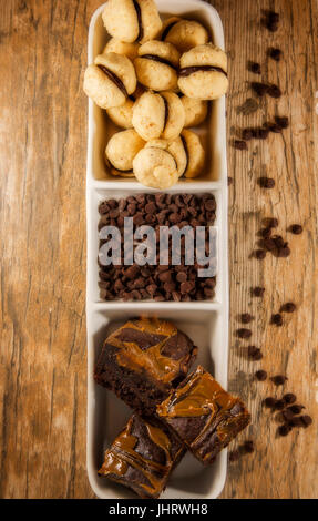 Bite size brownies and round mini chocolate cookies displayed in a white serving dish, with chocolate chips. Wooden background Stock Photo