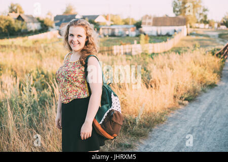 Portrait Of Young Pretty Plus Size Caucasian Happy Girl Woman With Blue Eyes, Wavy Brown Long Hair And Backpack In Summer Meadow Stock Photo