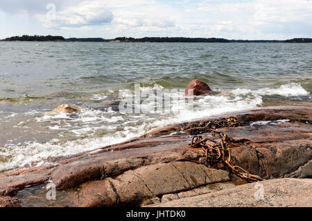 White foamy water waves hit into rocky seashore. Old rusty iron chain on rocks. Stock Photo