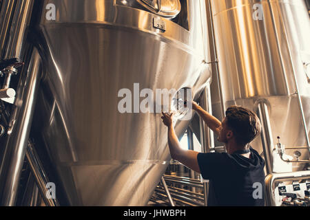 Young man working in beer manufacturing factory. Brewer working with industrial equipment at the brewery. Stock Photo