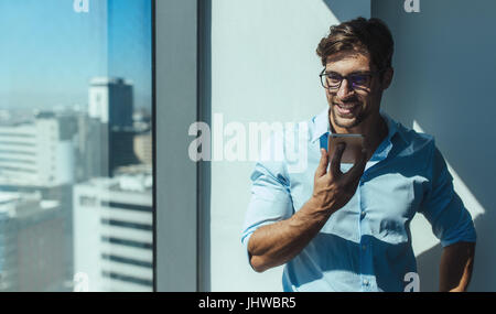 Businessman talking on speaker phone standing beside a window of highrise office building. Smiling young man with eyeglasses holding a mobile phone in Stock Photo